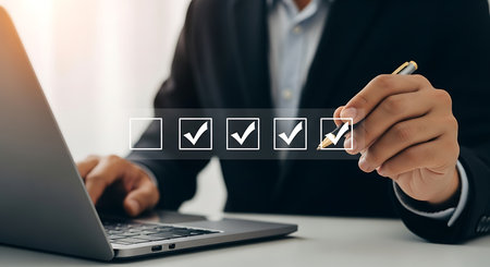 A person in a dark suit uses a pen to mark a checkmark on a digital checklist displayed on a screen, with a laptop in the foreground.の素材