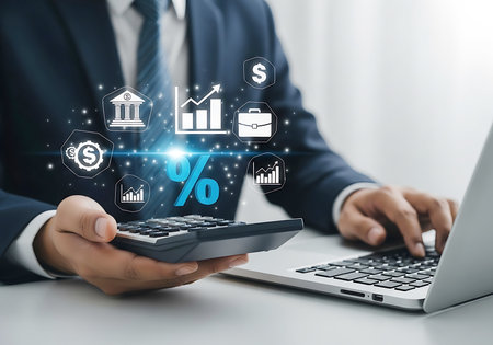 A man in a suit uses a calculator and laptop surrounded by glowing digital icons of finance graphs and currency.の素材