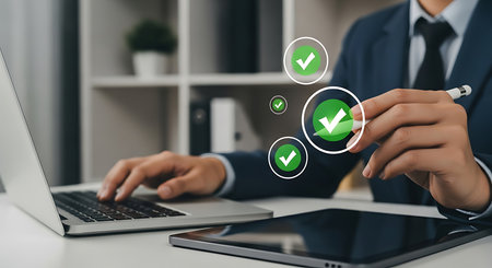 A man in a suit works on a laptop and tablet, with glowing green checkmarks overlaid on the scene.の素材