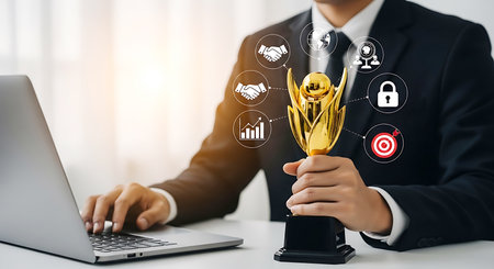 A man in a dark suit holds a golden trophy next to a laptop. Business icons float around the trophy.の素材