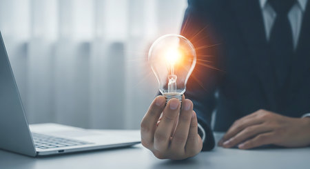 A person in a suit holds a glowing light bulb with a laptop nearby. The light bulb emits bright orange rays.の素材