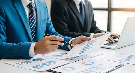 Two men in suits review financial charts and graphs on paper while one uses a laptop.の素材