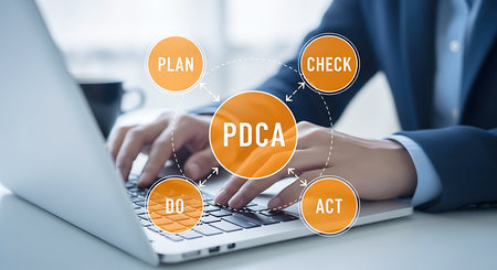 Hands typing on a laptop keyboard with an orange circular PDCA cycle diagram overlaid.の素材