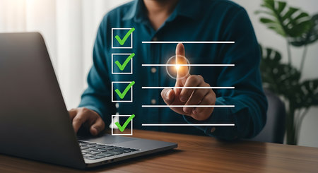 A person in a blue shirt touches a glowing circle on a digital checklist with green checkmarks next to completed tasks. A laptop is on a wooden desk.の素材