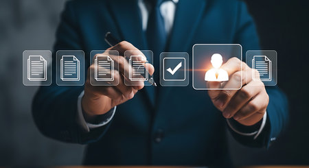 A man in a dark suit uses a pen to interact with a transparent digital screen displaying icons of documents, a checkmark, and a user profile.の素材