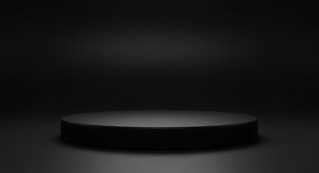 A dark circular podium with a matte finish sits on a dark studio floor, illuminated by subtle lighting creating soft shadows.の素材