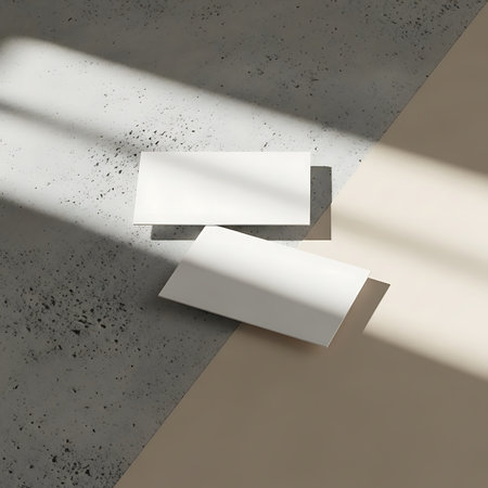 Two blank white rectangular cards rest on contrasting textured surfaces, one concrete grey with dark speckles, the other beige, casting soft shadows.の素材