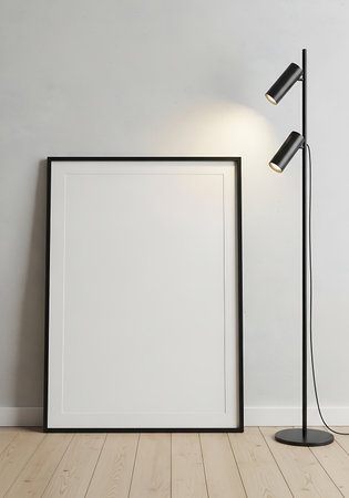 A large empty black framed poster leans against a textured white wall next to a modern black floor lamp with two spotlights.の素材