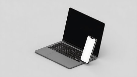 A silver laptop with a black screen sits next to a white smartphone on a plain gray surface.の素材