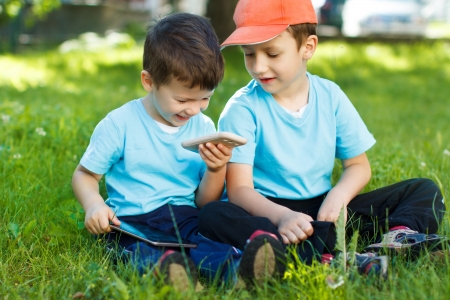 Happy young boys sitting in grass with wireless tablet and smartphoneの写真素材