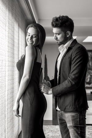 Stylish young man helps woman dressing up indoors, black and whiteの写真素材