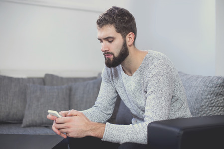 Young casual man messaging on smartphone on sofa in living roomの写真素材