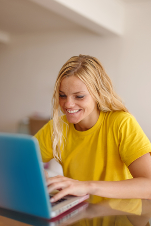 Happy young blonde woman in yellow t-shirt chatting on laptop, teeth smileの写真素材