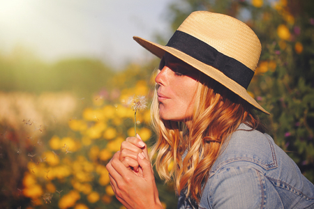 Young blonde woman in hat blowing dandelion at summer in natureの写真素材