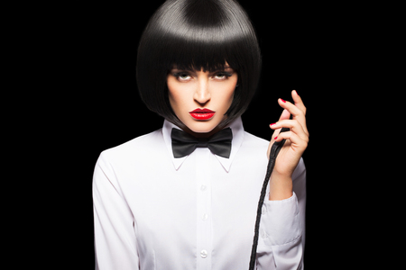 Sexy dominatrix in wig and red lips posing with whip, isolated on blackの写真素材