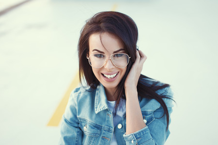 Happy young woman in jeans jacket and eyeglasses smiling outdoorの写真素材