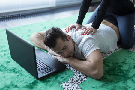 Young woman giving back rub to tired man lying on carpet with tabletの写真素材