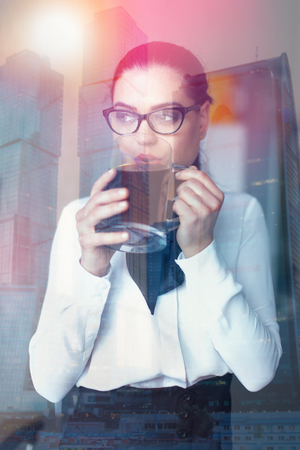 Young smart businesswoman in eyeglasses drinking big mug of coffee behind glasses in city, looking awayの写真素材