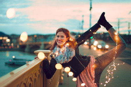 Happy young redhead woman doing yoga with fairy lights outdoors and smile, teal and orange style