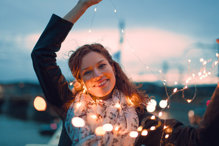 Happy young redhead woman playing with fairy lights outdoors and smile, teal and orange styleの写真素材