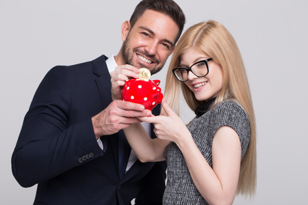 Happy young managers drop bitcoin into piggy bank on grey background portraitの写真素材