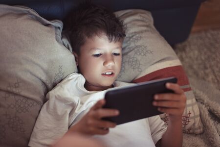 Little boy playing on digital tablet at night in dark bedroom