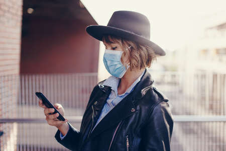 Young trendy woman in mask using smartphone outdoors in cityの写真素材