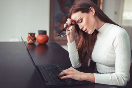 Young overloaded tired Caucasian woman has hadache at laptop in home office