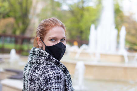 Young woman in black mask looking back at public park with fountain