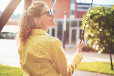 Happy young positive woman on swing in sunset, profile view, outdoors in park