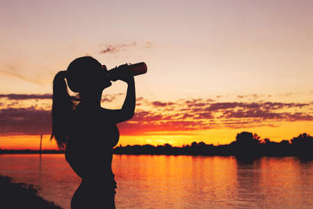 Tired thirsty woman silhouette drinking from bottle after workout in sunset