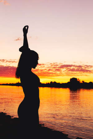 Sexy woman silhouette arms up in sunset at riverbank verticalの写真素材