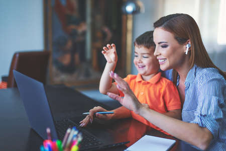 Young Caucasian mother with son waving at laptop in home office