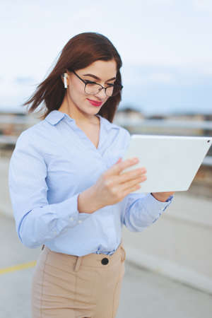 Young redhead 20s Caucasian businesswoman with hands-free headset using tablet outdoors