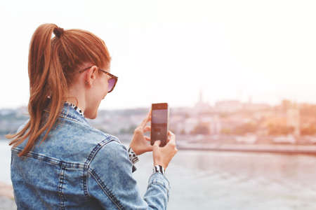 Young redhead woman taking picture of city panorama by smart phone in sunset, back view, horizontal