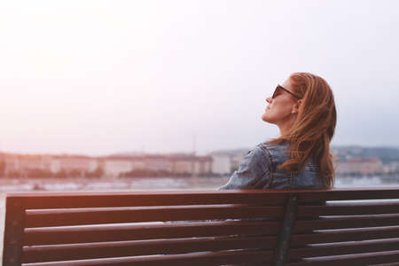 Young balanced redhead Caucasian woman sitting on bench at riverside meditating in sunlightの写真素材