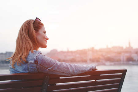 Young balanced redhead Caucasian woman sitting on bench resting at riverside in sunsetの写真素材