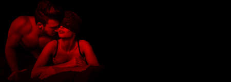 Sexy woman in lace eye cover in red light with young lover, foreplay in hotel roomの写真素材