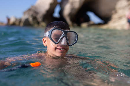 Happy young boy in goggles smiling in sea at rocky shore