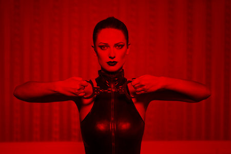 Sexy brunette woman in latex catsuit holding handcuffs in red light, bdsmの写真素材
