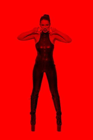 Sexy caucasian dominatrix in latex catsuit biting handcuffs in red lightの写真素材