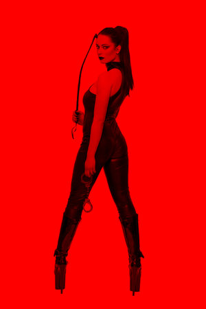 Sexy caucasian dominatrix in latex catsuit holding whip and turning back in red lightの写真素材