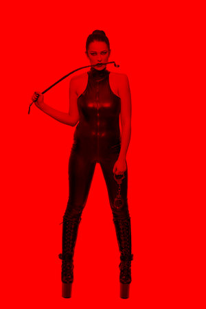 Sexy caucasian dominatrix in latex catsuit biting whip in red lightの写真素材