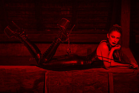 Sexy woman in catsuit and high heel platform boots laying on timber in red light, bdsmの写真素材