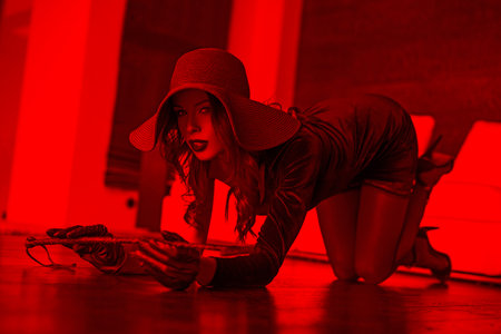 Sexy rich woman in hat and whip kneeling on floor indoor in red light, desireの写真素材