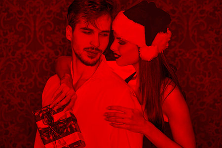 Young beautiful santa woman surprise boyfriend with gift in red light, embracing with present boxの写真素材