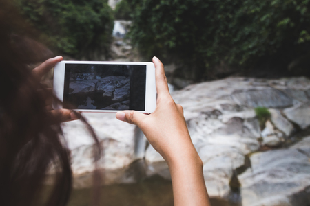 Asian woman tourist taking photo with camera phone at waterfall in Thailandの写真素材