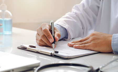 A male doctor with a uniform sits at a table in the doctor's office and writes a prescription on a piece of paper.の写真素材