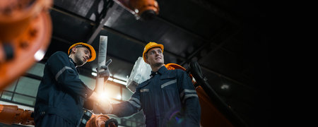 Male engineers wearing hard hats shaking hands in cooperation on project in machinery factory to...