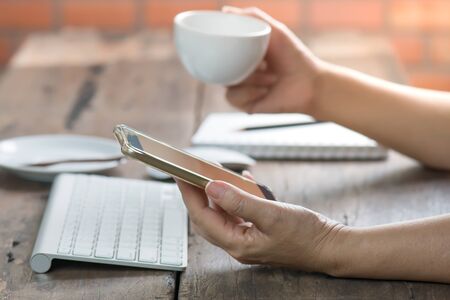 Woman working at old wooden table.Female hand holding smart phone with desktop computer keyboard...